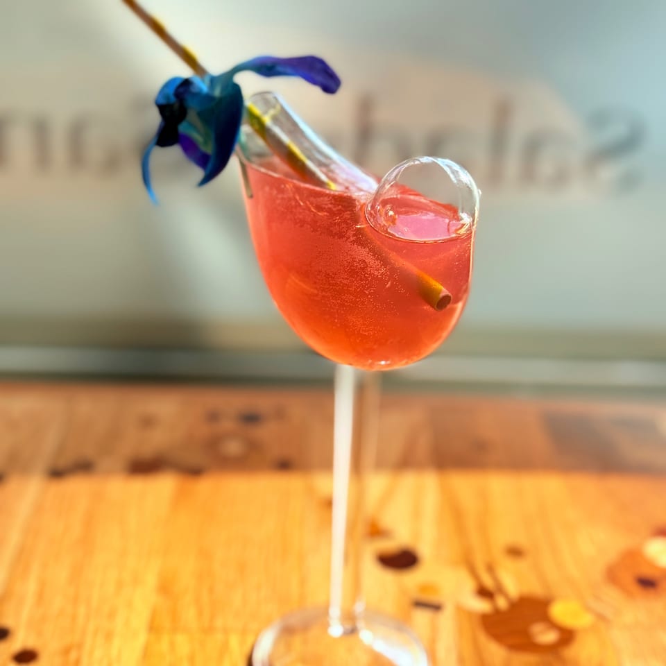 Bright pink cocktail in a stemmed glass, garnished with a blue flower and straw, sits on a sunlit wooden table.