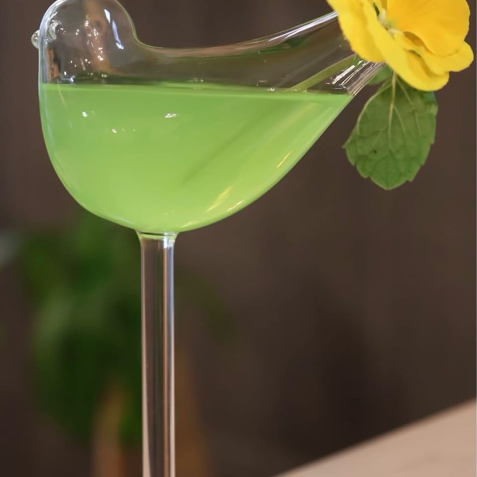 Bright green cocktail in an elegant stemmed glass, garnished with a yellow flower and leaf.