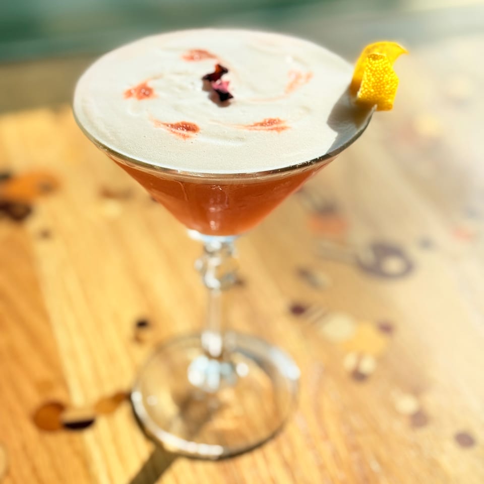 Frothy amber cocktail in a stemmed glass, garnished with citrus peel and bitters, sits on a sunlit wooden bar.