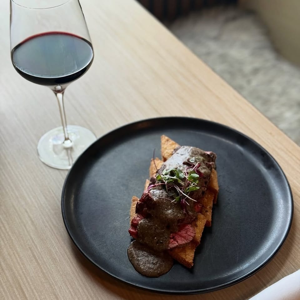 A black plate holds sliced steak over crisp toast with mushroom sauce and microgreens, accompanied by a glass of red wine.