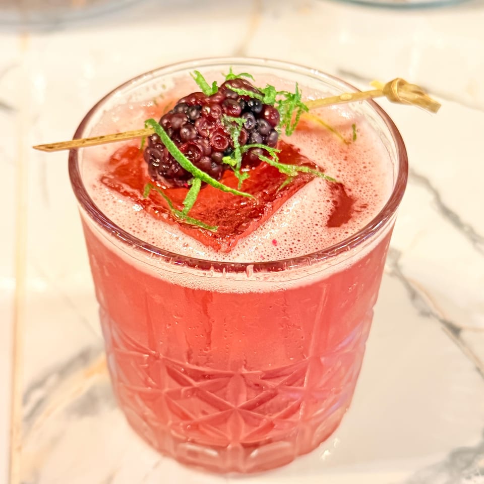 Pink cocktail in a cut-glass tumbler garnished with berries, lime zest, and a wooden skewer on a marble table.