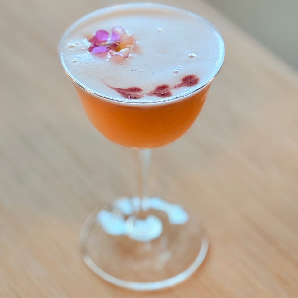 Orange cocktail in a coupe glass topped with frothy foam, edible flower petals, and red berry garnish on a wooden table.