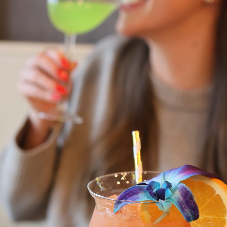 Woman sipping a green cocktail while an orange drink garnished with citrus and purple flowers sits in the foreground.