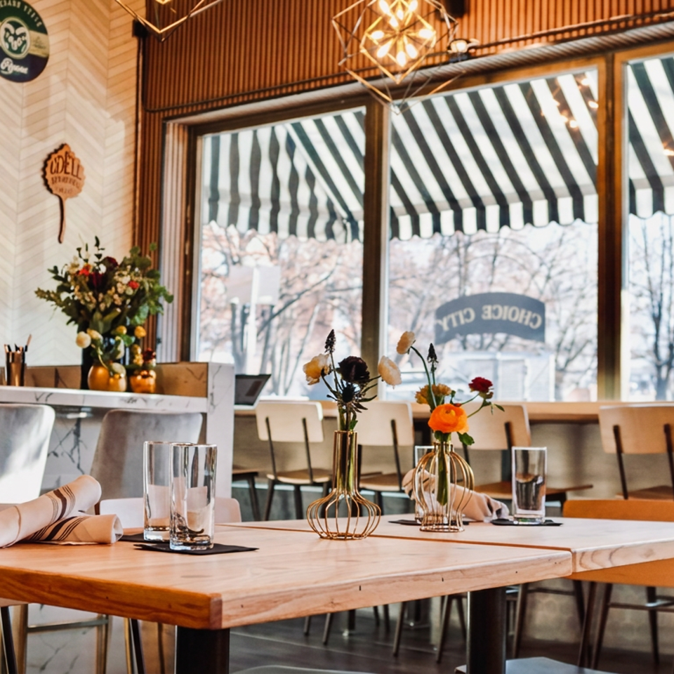 Warmly lit restaurant interior with set wooden tables, floral centerpieces, marble bar, beer décor, and large striped awning windows.