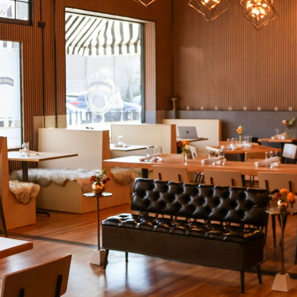 Warm, modern restaurant dining room with wood-paneled walls, set tables, leather sofa, floral accents, and pendant lighting.