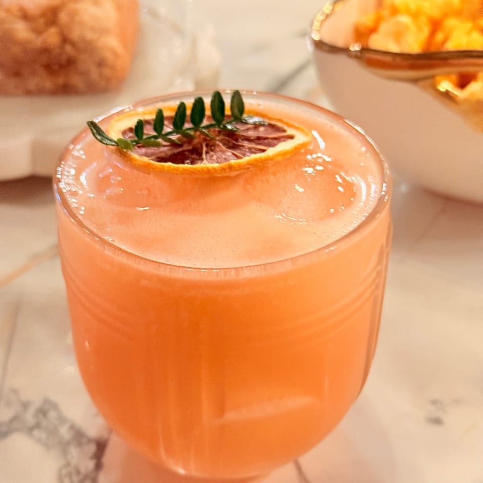 Frosty pink-orange cocktail garnished with a citrus slice and green herb sprig, served on a marble table with pastries behind.