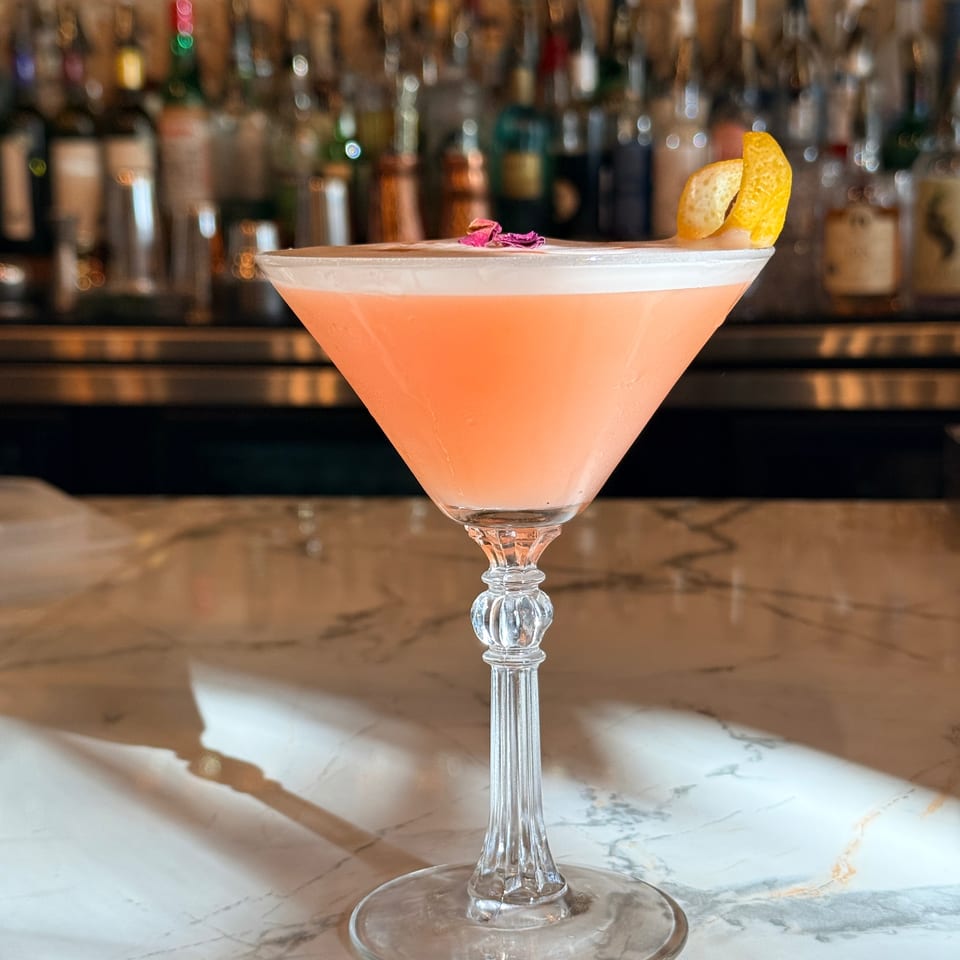 Pink cocktail in a coupe glass garnished with citrus peel and flower petals on a marble bar, with liquor bottles behind it.