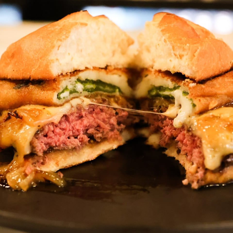 Cross-section of a cheeseburger on a bun, showing a juicy beef patty, melted cheese, caramelized onions, and green sauce.