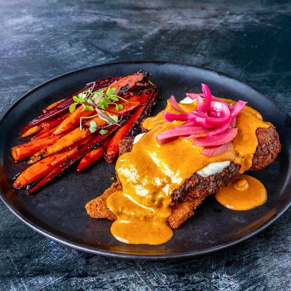Crispy fried cutlet topped with creamy orange sauce and pickled onions, served with glazed carrots on a black plate.