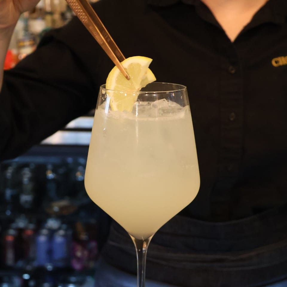 Bartender garnishes a pale citrus cocktail with a lemon wedge in a stemmed glass at the bar.