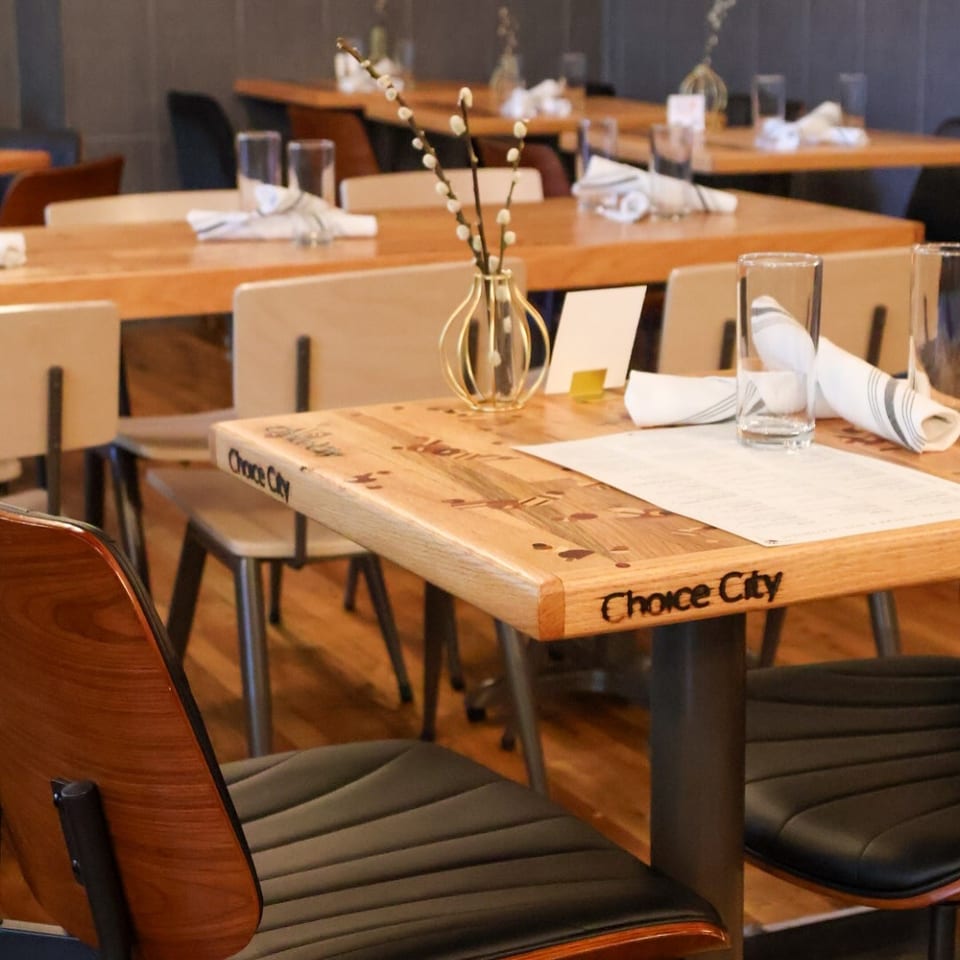 Warmly lit restaurant dining room with wooden tables, neatly folded napkins, water glasses, and decorative pussy willow branches.