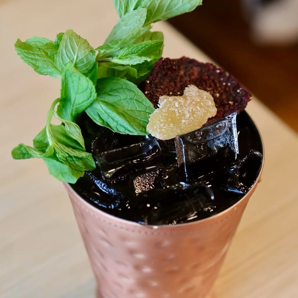 Copper mug filled with dark iced cocktail, garnished with fresh mint, crystallized ginger, and red fruit.